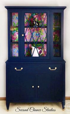 a blue china cabinet with flowers painted on the glass doors and drawers below it, in front of a white wall