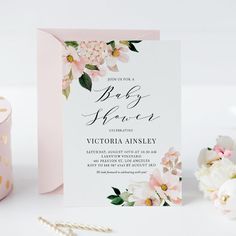 a pink and white floral baby shower is next to a candle, some flowers and a card