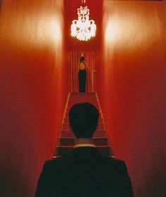 a man standing in front of a red wall next to stairs with chandelier