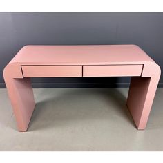 a pink desk sitting on top of a floor next to a gray wall with a black drawer underneath it