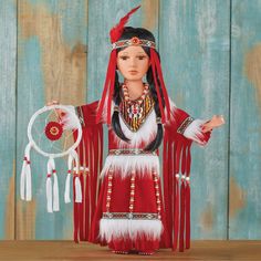 a doll dressed in red and white with feathers on her head, holding a dream catcher