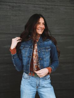 A woman with long dark brown hair standing in front of a wood wall wearing western fashion pieces great for spring including a medium wash jean jacket, wild rag, light wash blue jeans, and turquoise jewelry. Denim Vest For Workwear In Fall, Dark Wash Denim Vest With Pockets For Fall, Fall Denim Jacket With Flap Pockets, Winter Denim Jacket With Flap Pockets In Medium Wash, Fall Dark Wash Denim Vest With Pockets, Denim Vest For Work In Fall, Everyday Medium Wash Outerwear With Buttons, Fall Denim Jacket With Buttoned Pockets, Denim Outerwear With Flap Pockets In Medium Wash