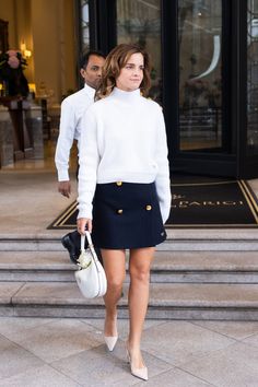 a woman in a white sweater and black skirt is walking down the street with her handbag