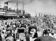 an old black and white photo of many people on the beach with ships in the background