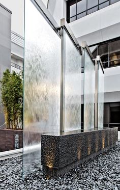 a large fountain in front of a building with glass walls and plants on the ground