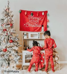 two young children in matching red pajamas playing with toys near a christmas tree and banner