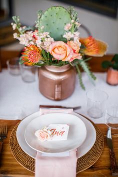 a place setting with pink napkins and flowers in a copper vase on the table