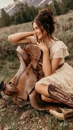 a woman sitting on the ground next to a brown horse wearing boots and a dress