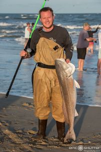 a man holding a fish on the beach with other people in the water behind him