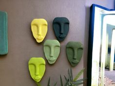 four different colored masks are on the wall next to a plant and mirror in front of it