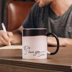 a man sitting at a table writing on a coffee mug with the words do i love you written on it