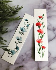 two bookmarks with watercolor flowers on them sitting on a marble table next to green leaves