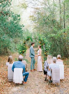 a couple getting married in the woods with their wedding guests sitting on white chairs and looking at each other