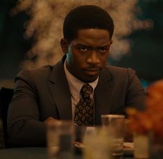 a man in a suit and tie sitting at a table with glasses on it, looking off to the side