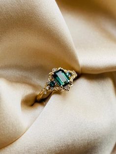 an emerald colored ring sitting on top of a white satin material with gold trimming