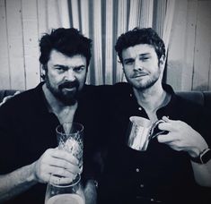 two men sitting next to each other holding drinks