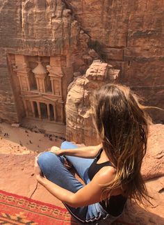 a woman sitting on the edge of a cliff in front of an ancient rock building