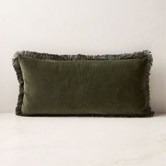 an olive green velvet pillow with fringe trim on the front and back, sitting on a white marble surface