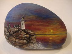 a painted rock with a lighthouse on it and the sun setting in the sky behind it