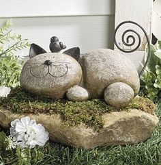 a cat statue sitting on top of a rock in the grass next to some flowers