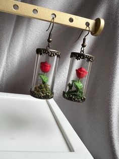 Add a magical touch to your fairytale style with new jewelry from Cosmic Created Magic! These beautiful detailed, delicate works of art are storybook inspired. The long stem red roses are made of clay. Resting below the rose is a bit of moss and greenery. The cylinder flower vase is made of glass, and the earrings hooks are bronze. You'll feel the beauty with these cool earrings. Elegant Rose Red Flower Earrings, Rose Red Flower Drop Earrings, Red Rose Earrings, Rose Red Flower-shaped Earrings For Pierced Ears, Vase Clay, Fairytale Style, Rose In A Glass, Rose Red Flower-shaped Earrings, La Rose