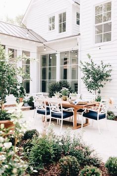 an outdoor dining table and chairs in front of a white house with potted plants