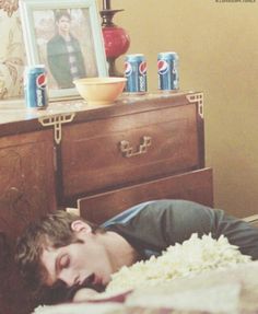 a young man laying in bed next to a dresser with two cans of soda on it