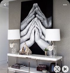 a table with two lamps and a painting on the wall behind it that has hands folded over each other