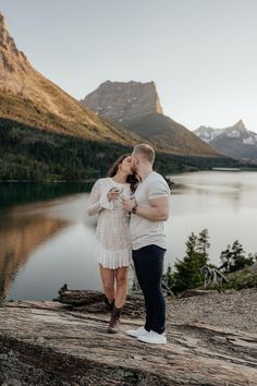 a man and woman standing next to each other on top of a rock near a lake