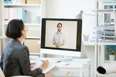 a woman sitting in front of a computer monitor with a man on it's screen