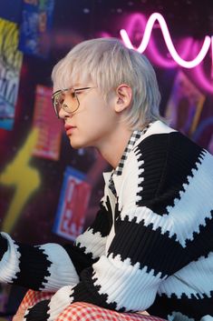 a man with white hair and glasses sitting in front of a neon sign holding a cell phone