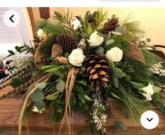 an arrangement of flowers and greenery on a table with pine cones, evergreens and white roses