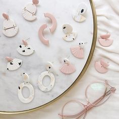 various pairs of pink and white earrings on top of a marble table next to eyeglasses