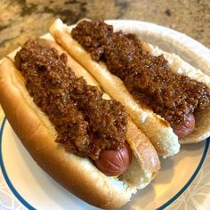 two hotdogs with chili on them sitting on a white and blue plate next to a marble counter