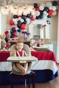 a young child sitting in a high chair at a table with red, white and blue balloons