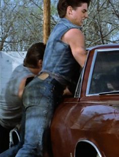 a young man leaning on the hood of a car with another person standing next to him