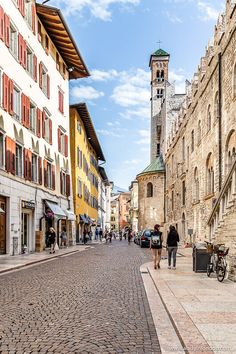 people are walking down the cobblestone street in an old european town with tall buildings