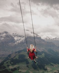 a woman in a red dress is swinging on a rope above the valley and mountains