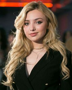 a woman with long blonde hair wearing a black suit and gold choker, posing for the camera