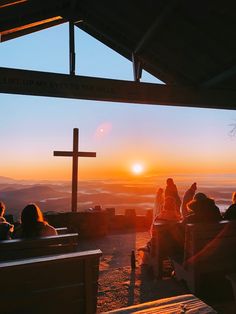 people sitting on benches watching the sun set over mountains and hills with a cross in the foreground