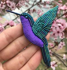 a hand holding a purple and green beaded hummingbird brooch with pink flowers in the background