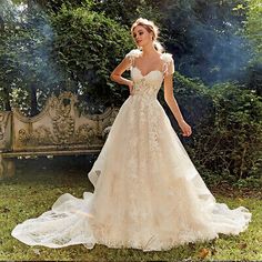 a woman in a wedding dress standing on the grass near a bench with trees and bushes behind her