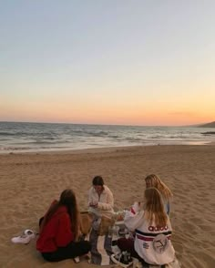 three people sitting on the beach at sunset eating sandwiches and drinking coffee while watching the sun set