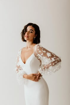 a woman wearing a white dress with sheer sleeves and flowers on the shoulders, standing in front of a white wall