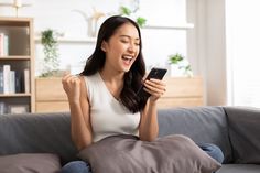 a woman sitting on a couch laughing and holding a cell phone in her hand while looking at the screen