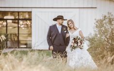 a bride and groom standing in front of a white barn with tall grass on the ground