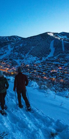 two people on skis in the snow near a town at night with lights lit up behind them