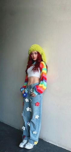 a woman with red hair wearing jeans and a rainbow sweater is standing against a wall