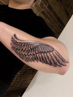 a man with a black and grey wing tattoo on his arm, next to a brick wall