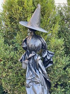 a statue with a black dress and hat on it's head, in front of some bushes